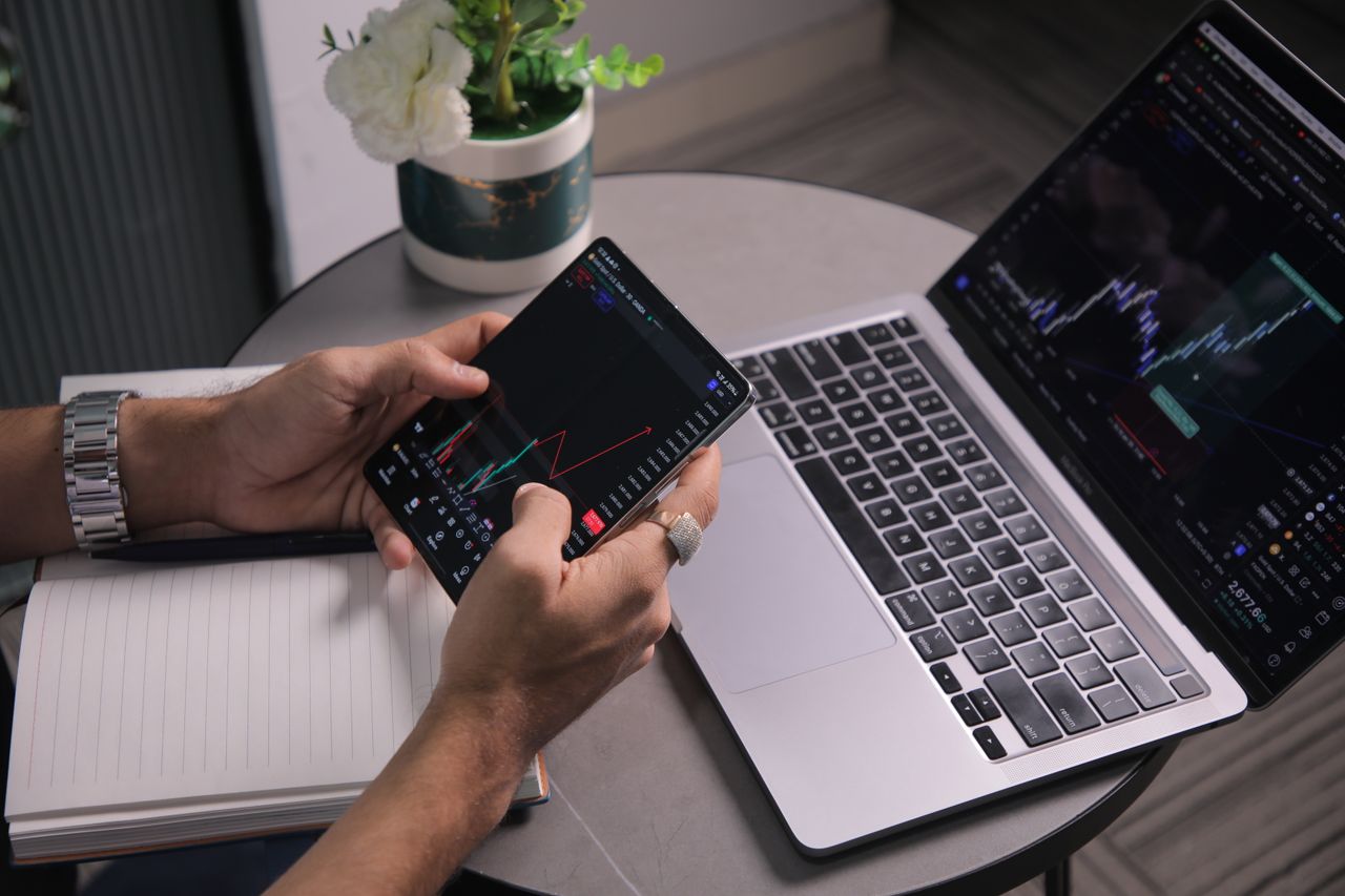 Man analyzing stock trends with laptop and mobile trading app at a modern workspace.
